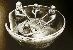 Piece of ceramic pottery with miniature of a scene during kiln firing, with figurines place around it two blowing the fire with long tubes and two attending the fuel